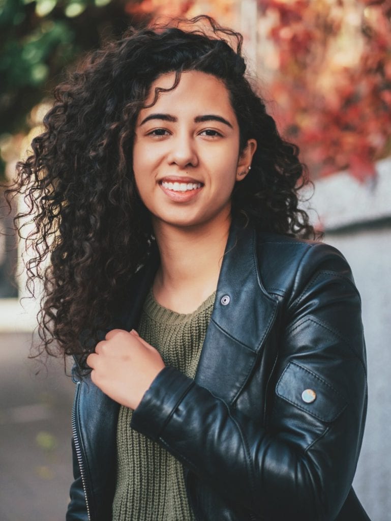 Portrait of happy young latino woman with beautiful curly hair in leather jacket and jeans standing in the street. Hispanic girl smiling. autumn season. Fall colors, park